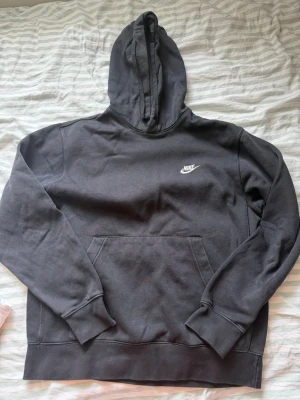 Svart Nike hoodie med logga - Klassisk svart hoodie från Nike med vit broderad logga på bröstet. Hoodien har en stor känguruficka framtill, ribbade muddar och en skön huva. Perfekt för en avslappnad och sportig stil. Bra bra skick. Storlek:M-L