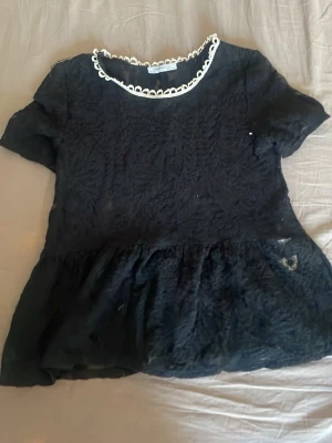 Svart spetsblus från Zara Trafaluc - Svart blus i spets från Zara Trafaluc med kort ärm och volang nertill. Rund halsringning med vit dekorativ kant. Blusen har ett feminint och trendigt uttryck med transparenta detaljer och blommönstrad struktur.