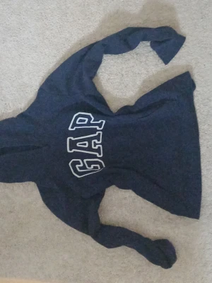 Mörkblå hoodie från GAP - Mörkblå hoodie från GAP med stor vit logga på bröstet. Klassisk modell med huva och långa ärmar. Perfekt för en avslappnad och cool stil. Mjuk och bekväm bomullsblandning.