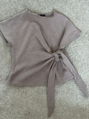 Beige topp med knyt från Zara - Trendig beige topp från Zara med korta ärmar och snygg knytning framtill. Toppen  är tillverkad i ett mjukt stickat material. Perfekt att styla med jeans eller kjol för en chill look.