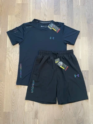 Svart träningsset från Under Armour - Snyggt svart träningsset från Under Armour med t-shirt och shorts. Setet har reflekterande loggor och textdetaljer i regnbågsfärger. Tillverkat i ett lätt och snabbtorkande material som passar perfekt för träning och sportaktiviteter. Hör av dig vid intresse, pris kan diskuteras!