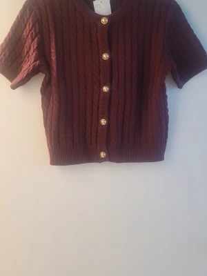 Vinröd kabelstickad kofta kort ärm - Kortärmad vinröd kabelstickad kofta med rund hals och guldfärgade knappar framtill. Ribbstickade muddar vid ärmslut och nederkant. Perfekt att styla över en topp eller klänning för en trendig look.