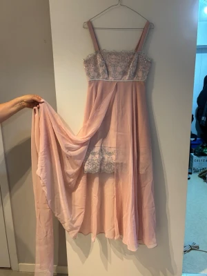 Lång rosa klänning med spetsdetaljer - Elegant långklänning i ljusrosa med smala axelband och vacker spetsdekoration över bysten och nedre delen. Klänningen har ett flödande, transparent tyg som ger ett drömmigt intryck och en extra tygdel som kan användas som sjal. Perfekt för dig som vill ha något romantiskt och unikt. Kommer med en scarf som man kan ha runt halsen. 