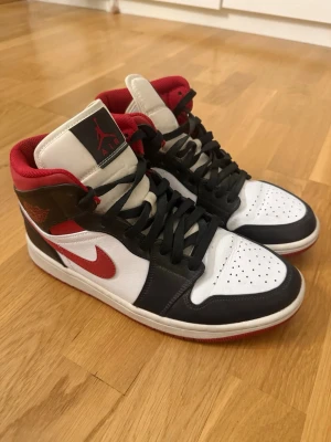 Air Jordan 1 Mid Svart/Vit/Röd - Säljer ett par Air Jordan 1 Mid sneakers från Nike i svart, vitt och rött. Skorna har klassisk hög siluett, svart läder på tå och sidor, vit tåbox och paneler samt röda detaljer på swoosh och krage. Ikonisk Jumpman-logga på plösen och vingar på sidan.