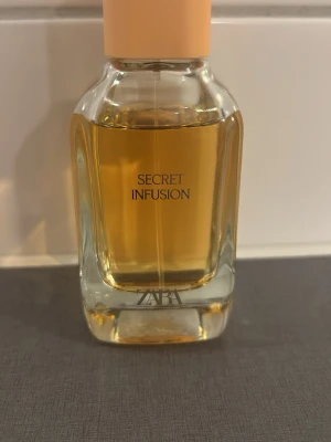 Zara Secret Infusion parfym - Zara Secret Infusion parfym i en stilren glasflaska med ljusbeige kork. Flaskan är transparent och visar den gyllene vätskan inuti. Perfekt för dig som gillar eleganta och moderna dofter.