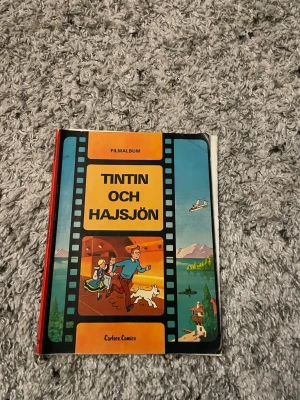 Tintin och Hajsjön filmalbum - Säljer boken 'Tintin och Hajsjön', ett filmalbum från Carlsen Comics. Omslaget är färgglatt med illustrationer av Tintin, Milou och andra karaktärer. Boken har hårda pärmar i kartong och är på svenska. Perfekt för dig som gillar klassiska äventyr och serier.