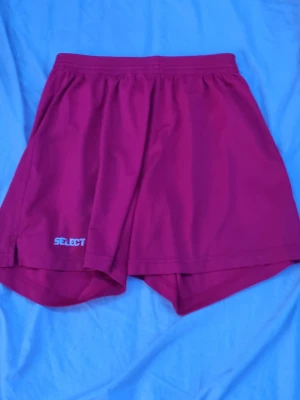 Röda sportshorts från Select - Röda sportiga shorts från Select med elastisk midja och logga på ena benet. Perfekta för träning eller fotboll, tillverkade i lätt och bekvämt polyestermaterial som andas. Klassisk passform och enkel design.