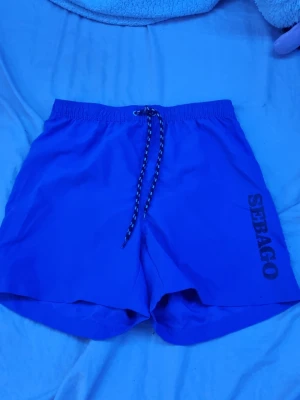 Blåa badbyxor från Sebago - Snygga blå badbyxor från Sebago med resår och dragsko i midjan. Tryckt logga längs ena benet och innerbyxa i mesh för extra komfort. Perfekta för stranden eller poolen.