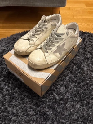Beige sneakers från Golden Goose - Snygga beige sneakers från Golden Goose med grå snörning och den ikoniska stjärnan på sidan i mocka. Skorna har en klassisk låg profil och är tillverkade i skinn med detaljer i mocka. Perfekta för en trendig och avslappnad stil. Pris kan diskuteras