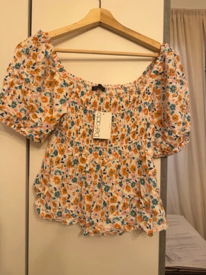 Blommig offshoulder topp från Defacto - Superfin blommig offshoulder topp från Defacto i vitt med orange, blå och rosa blommor. Toppen har korta puffärmar, smockad överdel och volangdetaljer nertill. Perfekt för en somrig look och lätt att matcha med jeans eller kjol.