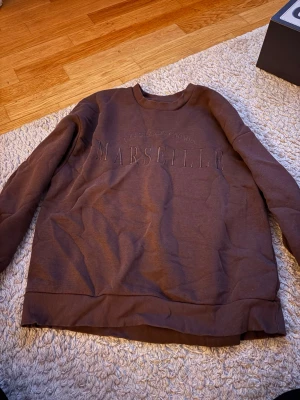 Brun sweatshirt från Gina Tricot - Säljer en brun sweatshirt från Gina Tricot med texten 'Marseille' broderad framtill. Tröjan har rund hals, ribbade muddar och är tillverkad i mjukt bomullsmaterial. Perfekt för en avslappnad och trendig look.