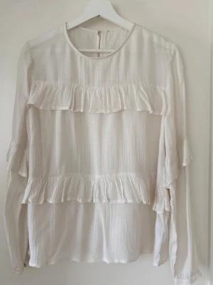 Vit blus med volanger från Soaked in Luxury - Vit blus med långa ärmar och volangdetaljer över bröstet och runt ärmsluten. Blusen har en rund halsringning och en diskret textur i tyget. Fina knappar i ärmslut och baktill.  Använd två gånger. Inga skavanker. Lite mer offwhite än vit.