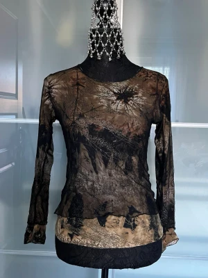 Mesh topp med batikmönster - Snygg mesh topp i transparent material med lång ärm och coolt batikmönster i svart och beige. Figurnära passform och rund halsringning, perfekt för att skapa en edgy look.
