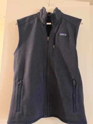 Mörkblå fleeceväst från Patagonia - Mysig mörkblå fleeceväst från Patagonia med hög krage och hel dragkedja. Västen har två sidofickor med dragkedja och en bröstficka med dragkedja. Perfekt lager-på-lager-plagg för kyliga dagar. Storlek small, pris kan diskuteras