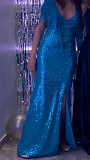 Blå balklänning glitter - Säljer en blåglittrig klänning med korsad rygg. Klänningen har en djup v-ringning och en hög slit till knäet. Endast använd en gång men är lite smutsig på innersidan av klänningen längst ner. Perfekt för bal eller fest.