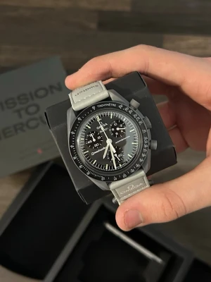 Omega x Swatch ”Mission to Mercury” - Oanvänd och i perfekt skick. Modellen har en stilren mörkgrå/svart färg med matta toner, inspirerad av planeten Merkurius. En diskret men samtidigt exklusiv look som passar till allt och ger en modern, clean känsla.  Designen är sportig men ändå elegant, vilket gör den lätt att bära både till vardags och vid mer uppklädda tillfällen. Lätt på handleden och mycket bekväm att använda.  Säljes då den inte kommer till användning. Snabba svar & skickas i