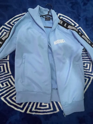 Ljusblå ziptröja från Hoodrich - Snygg ljusblå ziptröja från Hoodrich med svarta band och vit logga på ärmarna. Tröjan har ståkrage, dragkedja framtill och två fickor. Perfekt för en sportig och avslappnad stil.