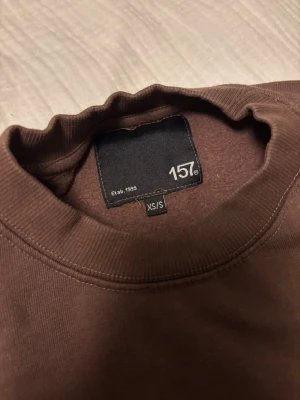 Brun sweatshirt från 157 XS/S - Mysig brun sweatshirt från 157 i storlek XS/S. Tröjan har rund halsringning, långa ärmar och ribbade muddar vid ärmslut och nederkant. Perfekt för en avslappnad och stilren look. Materialet känns mjukt och bekvämt mot huden.