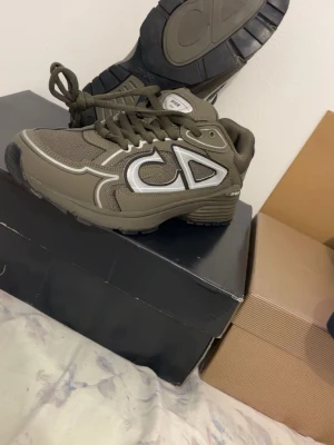 Olivgröna CD sneakers med silverdetaljer - Snygga olivgröna sneakers från CD med stora silverfärgade logodetaljer på sidan. Skorna har en chunky sula och meshpaneler för extra komfort och stil. Perfekta för dig som vill ha ett statement-piece till din outfit.