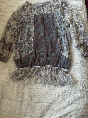Blommig transparent blus från Zara basic - Supersöt blus från Zara Basic i transparent material med blommigt mönster i lila, blått och beige samt paisleydetaljer framtill. Blusen har trekvartsärm, knappar vid halsen och volang nertill med dragsko. Perfekt för en trendig och romantisk look eller en varm fin sommar.