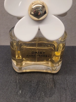 Daisy parfym från Marc Jacobs 100 ml. - Daisy från Marc Jacobs är en ikonisk parfym i en genomskinlig glasflaska med guldfärgad vätska. Flaskan har en unik kork dekorerad med stora vita blommor och gulddetaljer, vilket ger en lekfull och lyxig känsla. Perfekt för dig som gillar snygg design och fräscha dofter. MÄNGD SOM ÄR KVAR ÄR CA 60 ML.