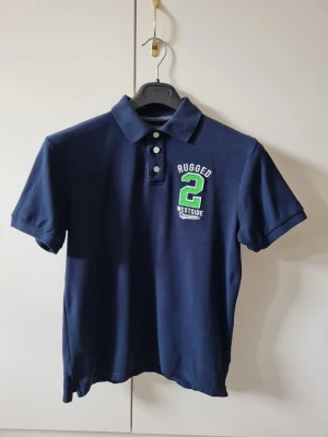 Pikétröja med tryck från Cubus strl 164 (barn) - Säljer en mörkblå pikétröja med krage och tre knappar framtill från Cubus. På bröstet finns ett broderat tryck med texten 'Rugged 2 Westside'. Material 100% bomull. Storlek 164. Kan hämtas i Vallentuna.