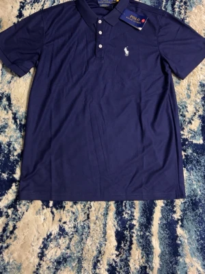 Mörkblå Polo Ralph Lauren pikétröja - Snygg mörkblå pikétröja från Polo Ralph Lauren med klassisk krage, tre knappar och den ikoniska vita logon broderad på bröstet. Tröjan är tillverkad i mjukt bomullsmaterial och har korta ärmar. Perfekt för en stilren och avslappnad look.