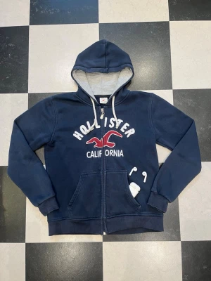 Marinblå zip-hoodie från Hollister - Snygg marinblå hoodie från Hollister med dragkedja, vit broderad text och röd logga på bröstet. Mjuk insida, stora fickor fram och justerbar huva med vita snören. Perfekt för chill dagar eller när du vill ha en casual look.