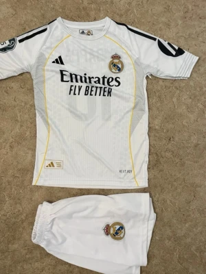 Real Madrid matchställ Mbappé 10 - Snyggt vitt Real Madrid matchställ från Adidas med Mbappé och nummer 10 på ryggen. Tröjan har svarta detaljer, klubbmärke och sponsortryck framtill samt gula linjer. Shortsen är vita med klubbmärke. Perfekt för fotbollsträning eller match.