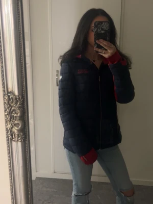 Mörkblå Tommy Hilfiger  pufferjacka med röd huva - Snygg mörkblå pufferjacka med röda detaljer och huva. Jackan har dragkedja framtill, två fickor med dragkedja och quiltad design. Perfekt för kalla vårdagar och riktigt skön att bära. Jackan har en sportig look och passar till många outfits.
