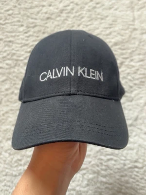 Svart keps från Calvin Klein - Snygg svart keps från Calvin Klein med vit broderad logga framtill. Klassisk böjd skärm och justerbar snapback-spänne bak för perfekt passform. Tillverkad i bomullstyg som känns både bekvämt och stilrent.