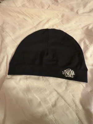 Svart mössa med broderad logga - Säljer en svart mössa i mjukt och stretchigt material med en vit broderad logga framtill. Enkel och stilren design som passar till det mesta. Perfekt för dig som gillar streetwear och vill ha en clean look.