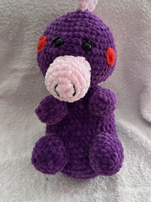Virkad lila drake - Supergullig handvirkad drake i mjukt lila garn med rosa mule och röda kinder. Svarta plastögon ger ett sött uttryck. Perfekt som dekoration eller present till någon som gillar unika mjukdjur. Materialet är fluffigt och mysigt polyester.