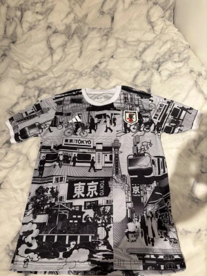 Japan Tokyo-inspirerad svartvit t-shirt adidas  - Unik t-shirt med svartvitt Tokyo-motiv och serietidningsstil. Trycket täcker hela tröjan med stadsmiljö, människor och japanska tecken. Rund halsringning och korta ärmar. Perfekt för dig som gillar streetwear och grafiska prints.