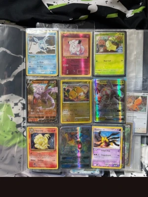 Pokémonkort samling i pärm - Stor samling Pokémonkort i plastfickor, med flera holografiska och färgglada kort. Blandade motiv med olika Pokémon, både klassiska och nyare. Kort i olika färger och stilar, vissa med glansig yta. Perfekt för samlare eller Pokémonfans.