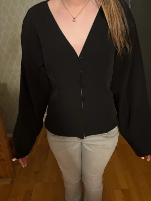 Svart blus med v-ringning och knappar - Stilren svart blus med djup v-ringning och knäppning framtill. Blusen har långa, vida ärmar och en rak passform som ger en avslappnad men trendig look. Perfekt att matcha med jeans eller kjol för en snygg outfit.