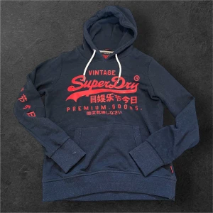  Fet Superdry hoodie - Snygg mörkblå hoodie från Superdry med röd logga och japanska tecken på bröstet och ärmen. Klassisk känguruficka, vit snörning i huvan och mjukt material. Perfekt för dig som gillar streetstyle och vill sticka ut med coola detaljer.
