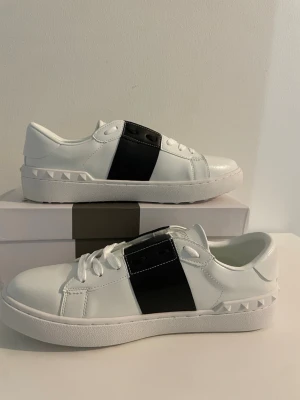 Vita Valentino Open Garavani Sneakers Storlek 43 - Snygga vita sneakers från Valentino med en bred svart läderdetalj över snörningen och coola nitar längs hälen. Skorna har en klassisk låg profil och är tillverkade i slätt läder med vita skosnören och tjock vit sula. Perfekta skor för dig som vill ha nått som passar till allt🤍 