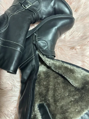 Svarta fodrade boots med spänne - Snygga svarta boots med mjukt foder och dragkedja på sidan. Dekorativa spännen och vita kontrastsömmar ger en cool look. Perfekta för kalla dagar när du vill hålla stilen och värmen. Materialet är konstläder och insidan är pälsfodrad.