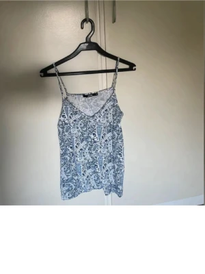Vit och blå paisleymönstrad linnetopp - Snygg linnetopp med tunna axelband och paisleymönster i vitt och blått. Toppen har v-ringning framtill och är perfekt för varma dagar. Lätt och luftig känsla, passar till jeans eller kjol.