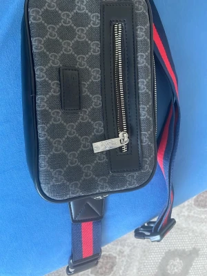 Svart axelväska med monogrammönster - Snygg svart axelväska från Björn Borg med grått monogrammönster och en silverfärgad dragkedja framtill. Väskan har en justerbar axelrem i blått och rött samt detaljer i konstläder. Perfekt för dig som vill ha en trendig och praktisk väska.