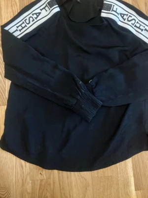 Svart hoodie med logga på ärmarna - Svart hoodie från Vero Moda med vita och svarta loggor längs båda ärmarna. Klassisk huva och ribbade muddar vid ärmsluten. Perfekt för en avslappnad och trendig look. Materialet känns mjukt och bekvämt.