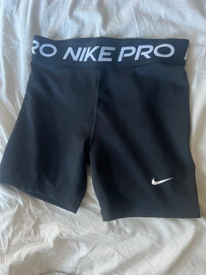 Svarta Nike Pro shorts - Svarta Nike Pro shorts med bred resår i midjan där det står 'NIKE PRO' i vitt. Tight passform och tillverkade i ett stretchigt syntetmaterial, perfekta för träning eller sport.