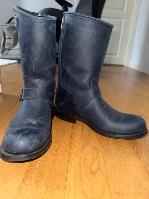 Svarta läderstövlar med spänne - Snygga svarta stövlar i läder med högt skaft och coolt spänne vid ankeln. Klassisk modell med rund tå och robust sula, perfekta för dig som gillar en stilren och tidlös look. Dragkedja baktill för enkel påtagning.