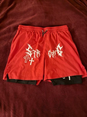 Röda shorts med tryck och snörning - Säljer ett par röda shorts med vita grafiska tryck och korsdetaljer framtill. Shortsen har svart innerbyxa och snörning i midjan för extra komfort. Perfekta för dig som gillar streetwear och vill sticka ut.