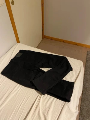 Svarta jeans med rak passform - Säljer ett par svarta jeans med klassisk rak passform. Jeansen har fem fickor och en läderpatch på linningen. Materialet är denim och färgen är djup svart, perfekt för en stilren look.