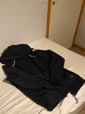 Svart hoodie från Björn Borg - Säljer en svart hoodie från Björn Borg med dragkedja och huva. Hoodien har ribbade muddar och en patch med logotypen på ena fickan. Perfekt för chill dagar och har en klassisk design med mjukt material.