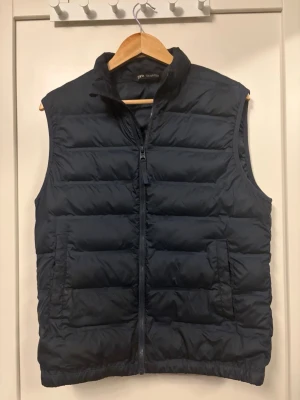 Mörkblå dunväst från Zara - Snygg mörkblå dunväst från Zara med hög krage och dragkedja framtill. Västen har två praktiska fickor med dragkedja och quiltad design som ger en sportig look. Perfekt att slänga över en hoodie eller tröja för extra värme.