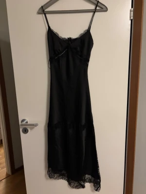 Svart klänning med spetsdetaljer - Elegant svart klänning med tunna axelband och v-ringning. Tillverkat i ett satinliknande material och dekorerat med spets vid byst och nederkant. Klänningen har en figurnära passform. I storlek 36/S från Gina Tricot. Helt ny med prislappkvar , aldrig använd. 
