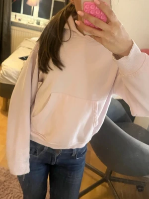 Ljusrosa hoodie med magficka - En enkel och snygg ljusrosa hoodie med huva och stor magficka framtill. Hoodien har ribbade muddar vid ärmslut och nederkant, samt en avslappnad passform. Perfekt för en chill och bekväm stil.